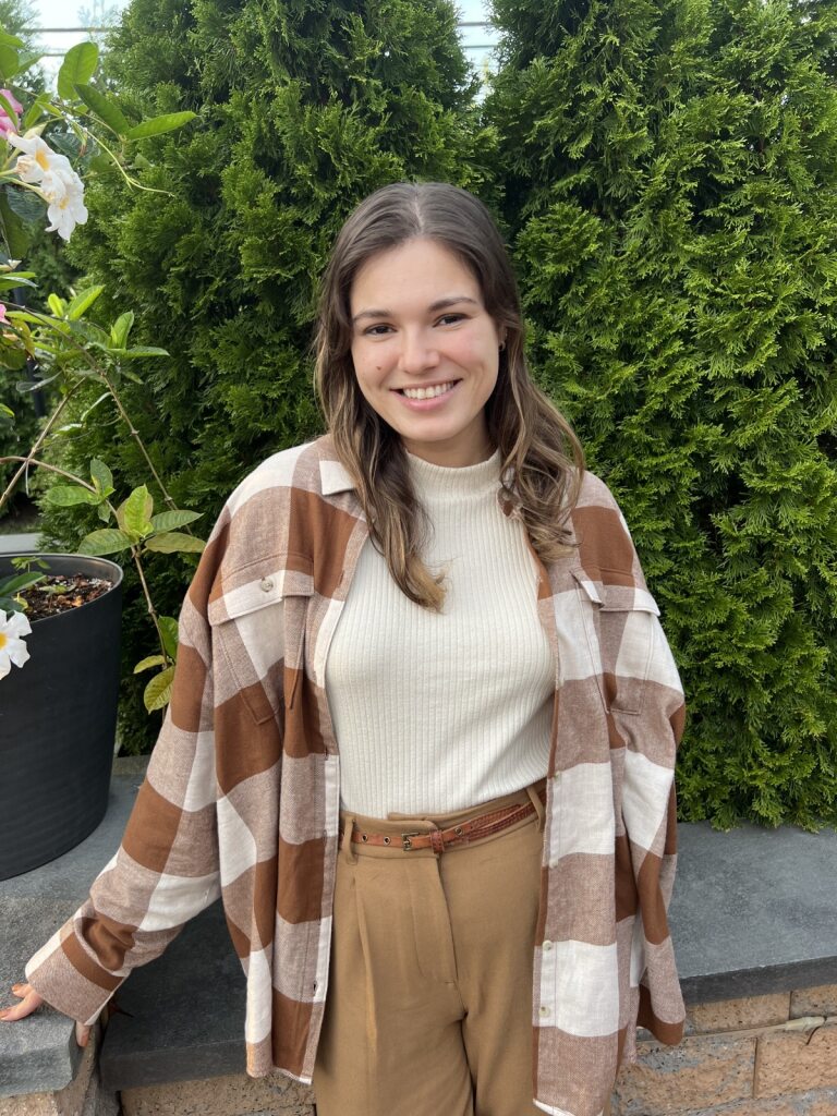 Samantha Blaine, MD "A person wearing a brown shirt standing outdoors on a mountain trail, with a view of a forested valley and rocky cliffs in the background."