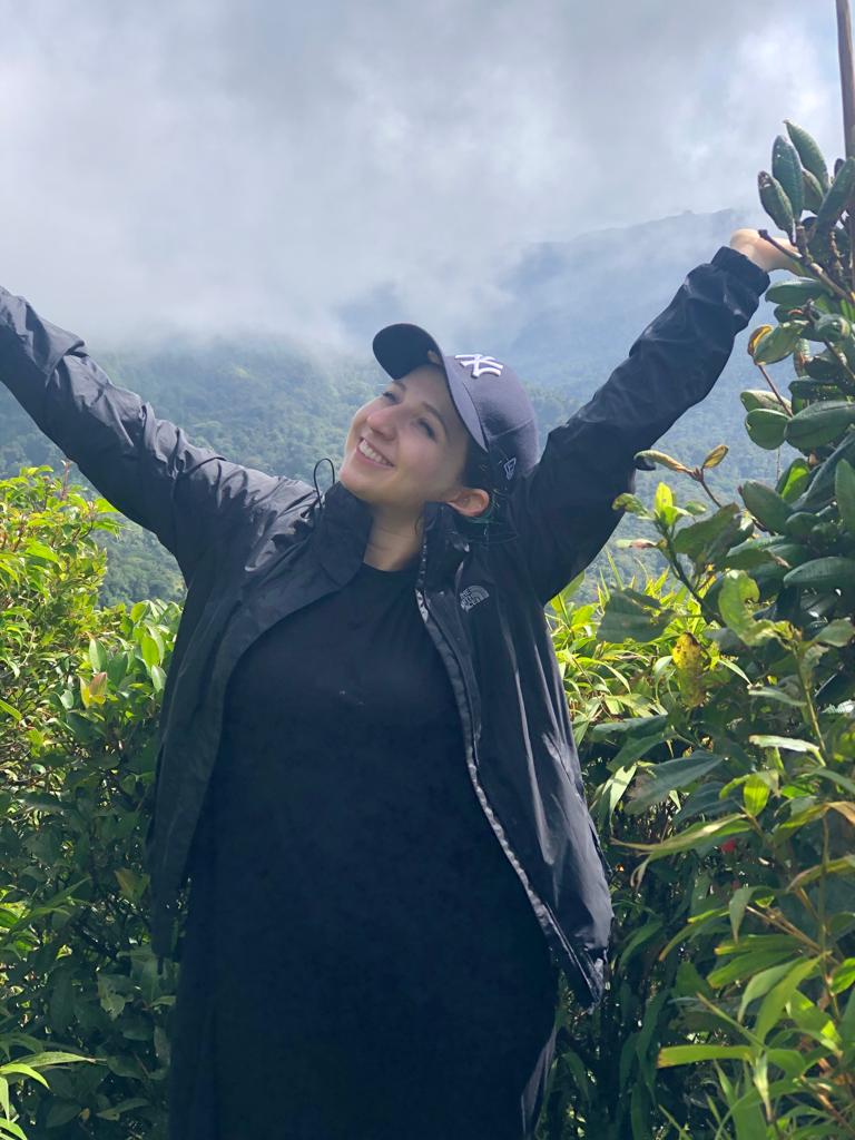 Leah Gancz, MD "Person standing outdoors with arms raised in front of green foliage and misty mountains."