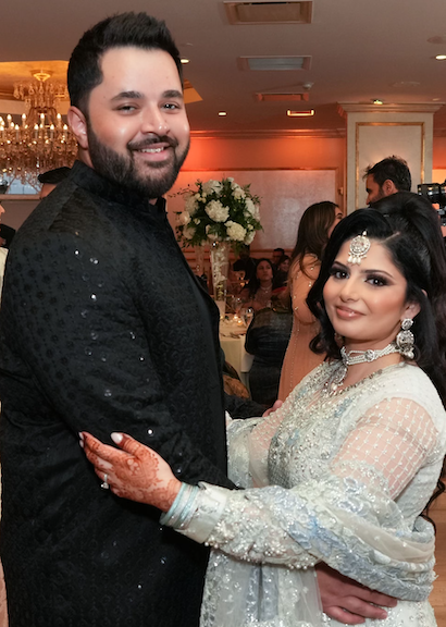 Zaid Ahmed, DO "A man and a woman standing together at an indoor event, looking at the camera. The man is wearing a black traditional outfit, and the woman is wearing a light-colored embroidered outfit with jewelry. The background shows a decorated venue with warm lighting and other people present."