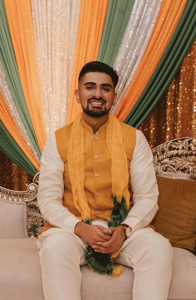 Asim Shahid, DO "Person sitting on a couch in front of a backdrop with green, orange, and white draped fabric, wearing traditional attire and holding green leaves."
