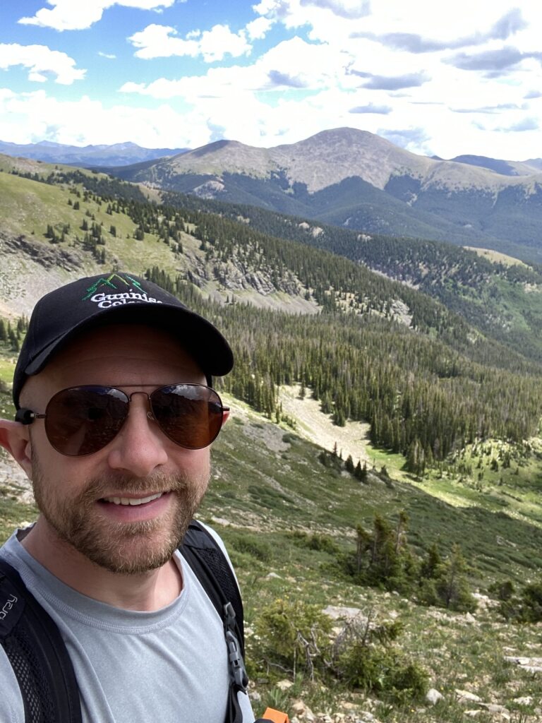 Cory Emborski, MD "Person taking a selfie outdoors with mountains, trees, and a partly cloudy sky in the background."