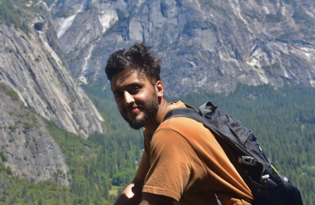 Danyaal Ain, MD "A person wearing a brown shirt and a backpack standing on a mountainside, looking toward the camera. The background shows a forested valley and steep rocky cliffs under clear daylight."