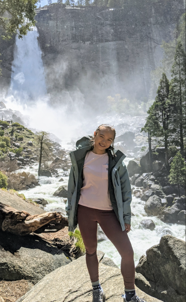 Jin Yao Li, DO "Person standing on rocky terrain in front of a large waterfall surrounded by greenery and tall trees."