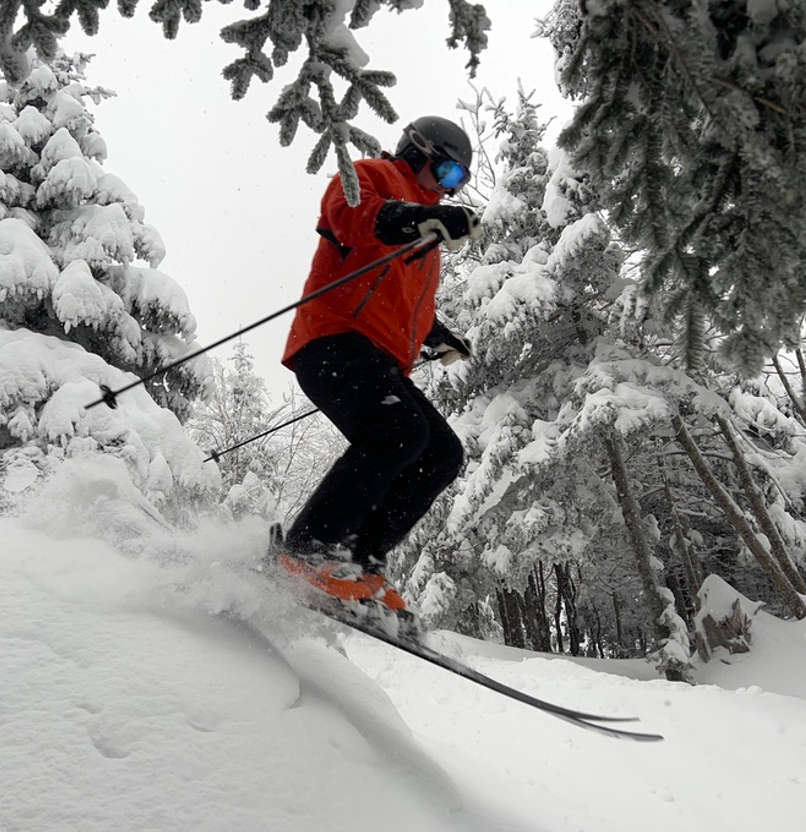 Andrew Mozier, DO "Person skiing downhill in a snowy forest, wearing a red jacket, black pants, and a helmet with goggles."