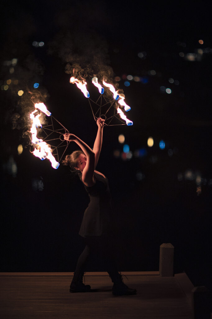 Laura Tran, MD "Person performing a fire dance at night, holding two flaming objects with blurred lights in the background."