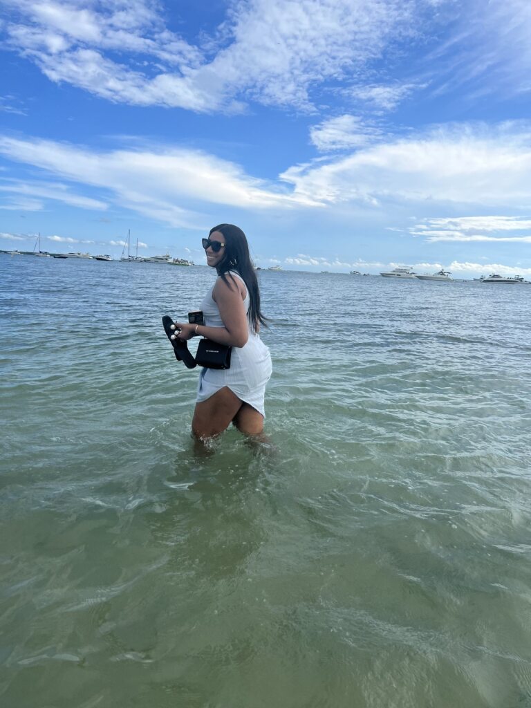 Chinenye Okudoh, MD "Person standing in shallow ocean water with a partly cloudy sky and distant buildings in the background.