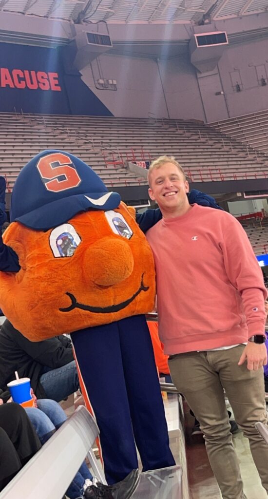 Justin Townsend, DO "Person standing next to a large mascot with an orange head and blue hat featuring the letter 'S', in an indoor stadium with seats and people in the background."