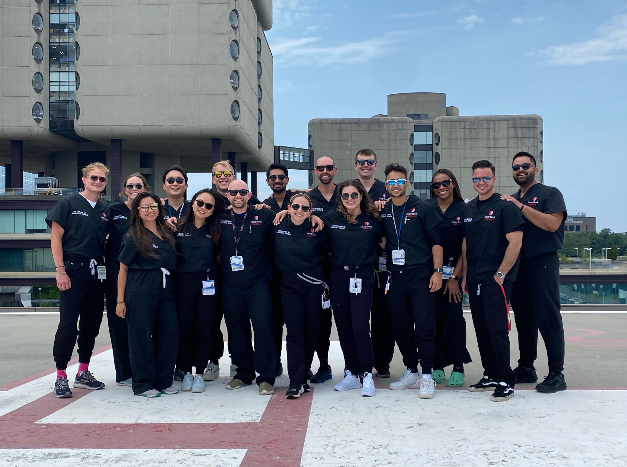 Class of 2026 "A group of sixteen people standing closely together on a rooftop helipad, all wearing matching black uniforms with ID badges and lanyards. They are smiling and posing for a group photo under a clear blue sky, with tall concrete buildings visible in the background. The group appears to be part of a professional or medical team."