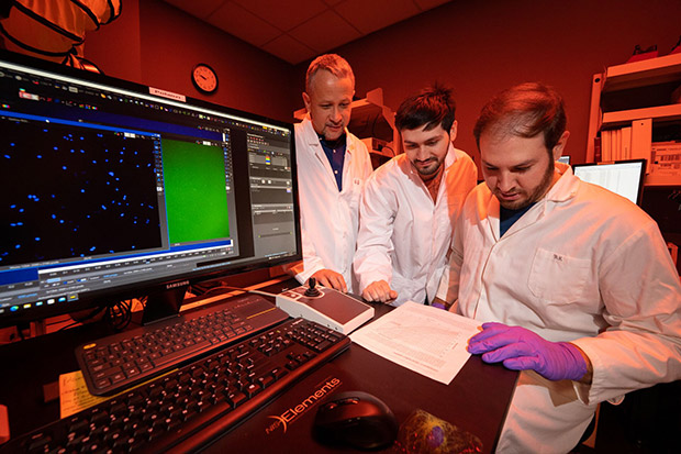 Three scientists in lab coats analyzing data on a computer screen in a dimly lit laboratory