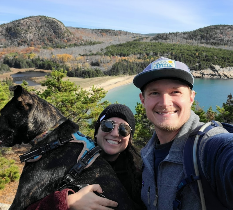 Ian Mathies, MD "Two individuals and a dog posing for a selfie outdoors with hills, trees, and a body of water in the background.