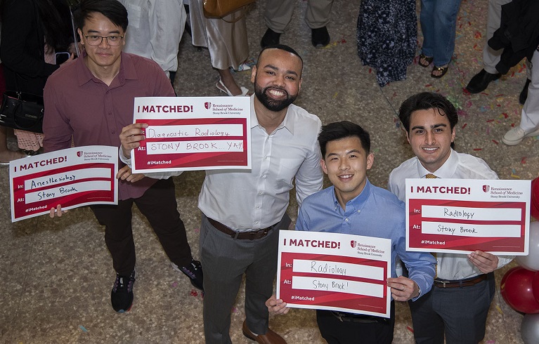 Four medical students celebrating Match Day, looking up at the camera and holding red-and-white 'I MATCHED!' signs showing specialties such as Anesthesiology and Diagnostic/General Radiology and 'Stony Brook' as their residency site