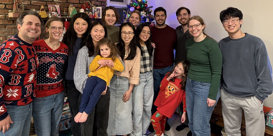 Rahme Lab members gathered in front of a decorated Christmas tree and fireplace, including two young children.